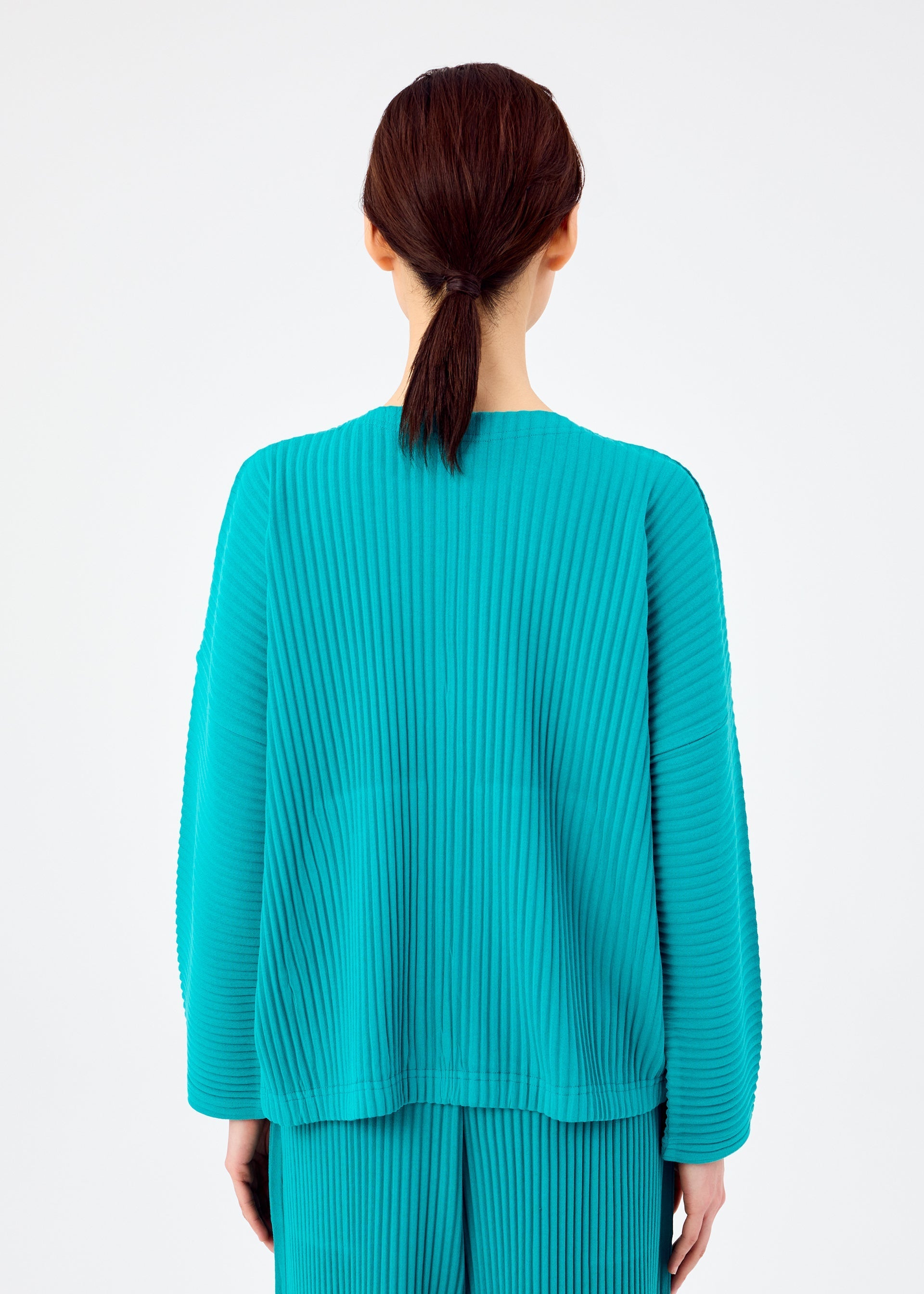 Zoomed view A person with dark brown hair tied back is seen from behind, wearing the bright teal SWEATS PLEATS long-sleeved pleated cardigan and matching pleated pants by me ISSEY MIYAKE, against a plain white background.