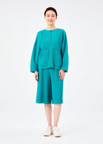 A person stands facing forward, dressed in the SWEATS PLEATS cardigan and matching knee-length shorts by me ISSEY MIYAKE, paired with white slip-on shoes. The background is plain white.