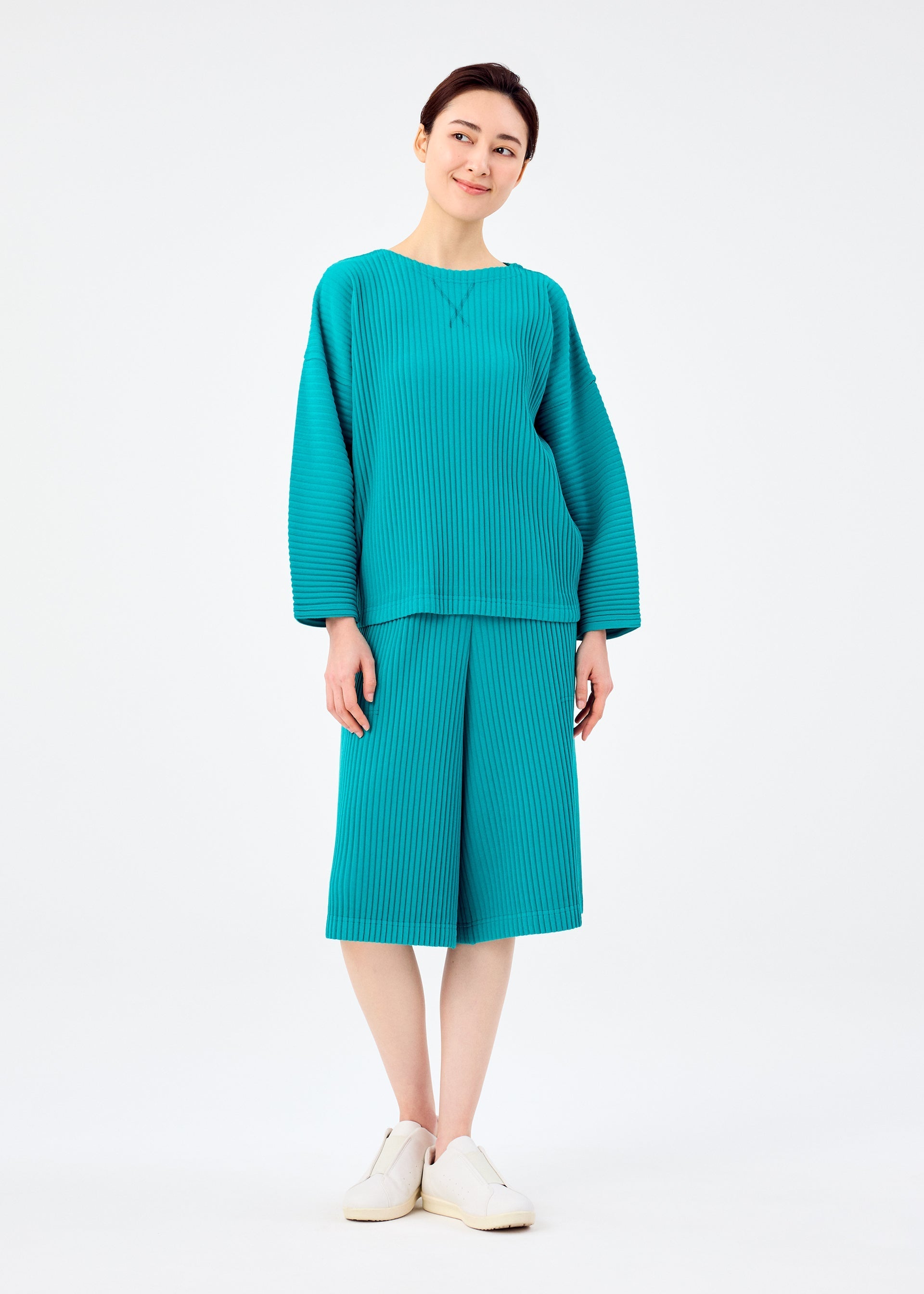 Zoomed view A person stands and smiles in a loose-fitting teal SWEATS PLEATS top with matching knee-length jersey fleece shorts by me ISSEY MIYAKE and white slip-on shoes, set against a plain white background.
