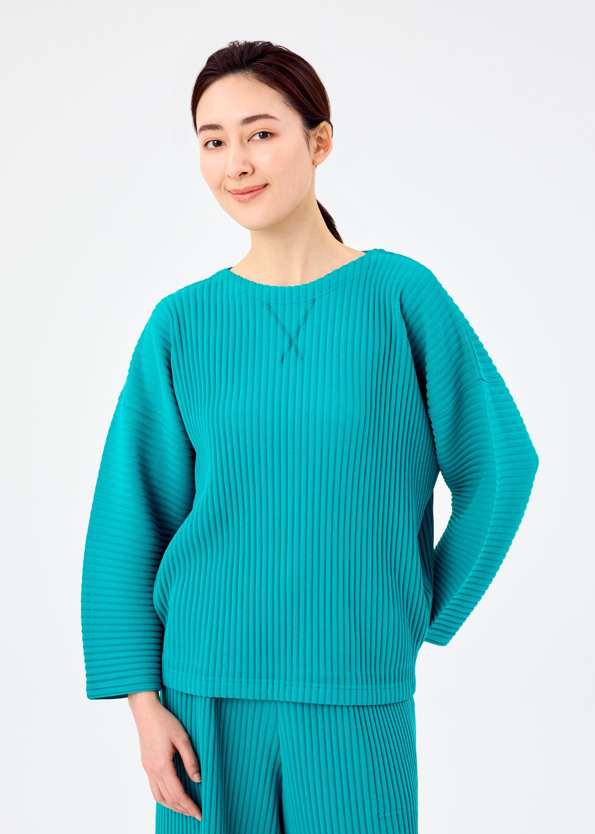 Zoomed view A woman with dark hair tied back wears the loose teal SWEATS PLEATS set by me ISSEY MIYAKE, standing against a white background and slightly smiling with one hand behind her back.