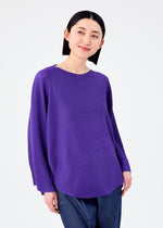 A person with short black hair models the me ISSEY MIYAKE CURVE KNIT STRETCH PLEATS shirt in purple and blue jeans, standing against a plain white background and smiling slightly while looking to the side.