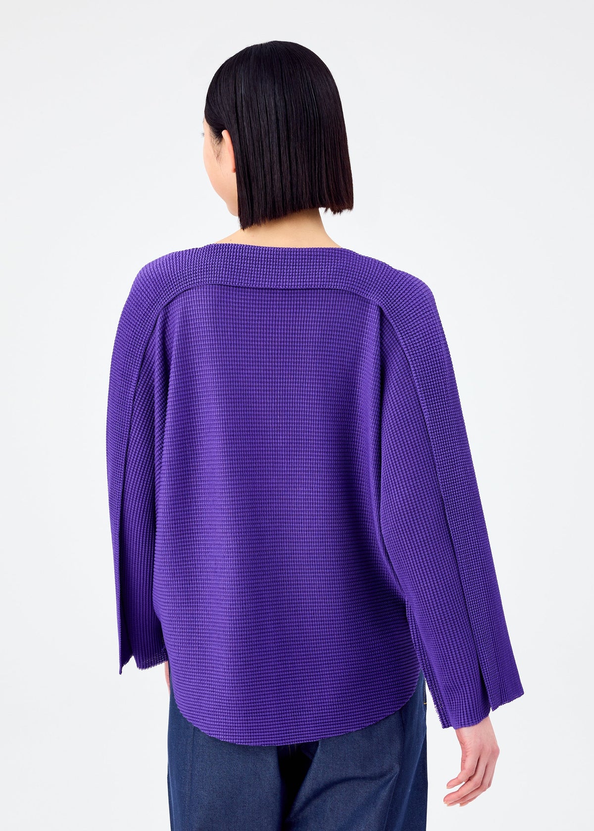 A person with straight, chin-length dark hair is seen from the back wearing the me ISSEY MIYAKE CURVE KNIT STRETCH PLEATS, a loose, textured purple shirt with wide sleeves and dark pants, set against a plain white background. view 7 of 8