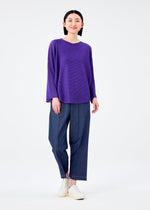 A person stands against a plain white background, smiling gently with one foot forward, wearing the CURVE KNIT STRETCH PLEATS shirt in purple by me ISSEY MIYAKE, blue wide-leg pants, and white slip-on shoes.