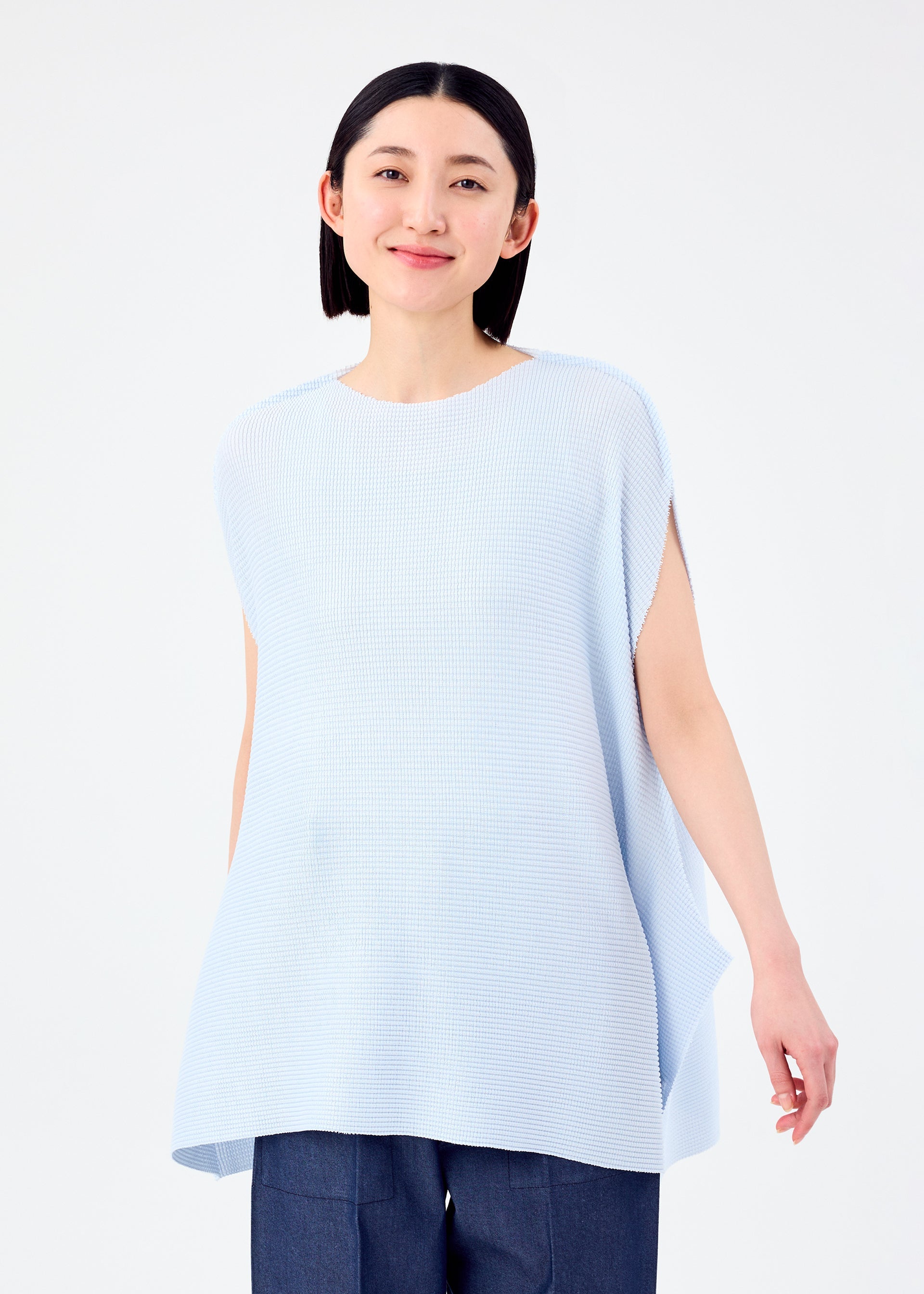 Zoomed view A person with straight black hair smiles gently, wearing the me ISSEY MIYAKE CURVE KNIT STRETCH PLEATS top in light blue and dark pants, standing against a plain white background.