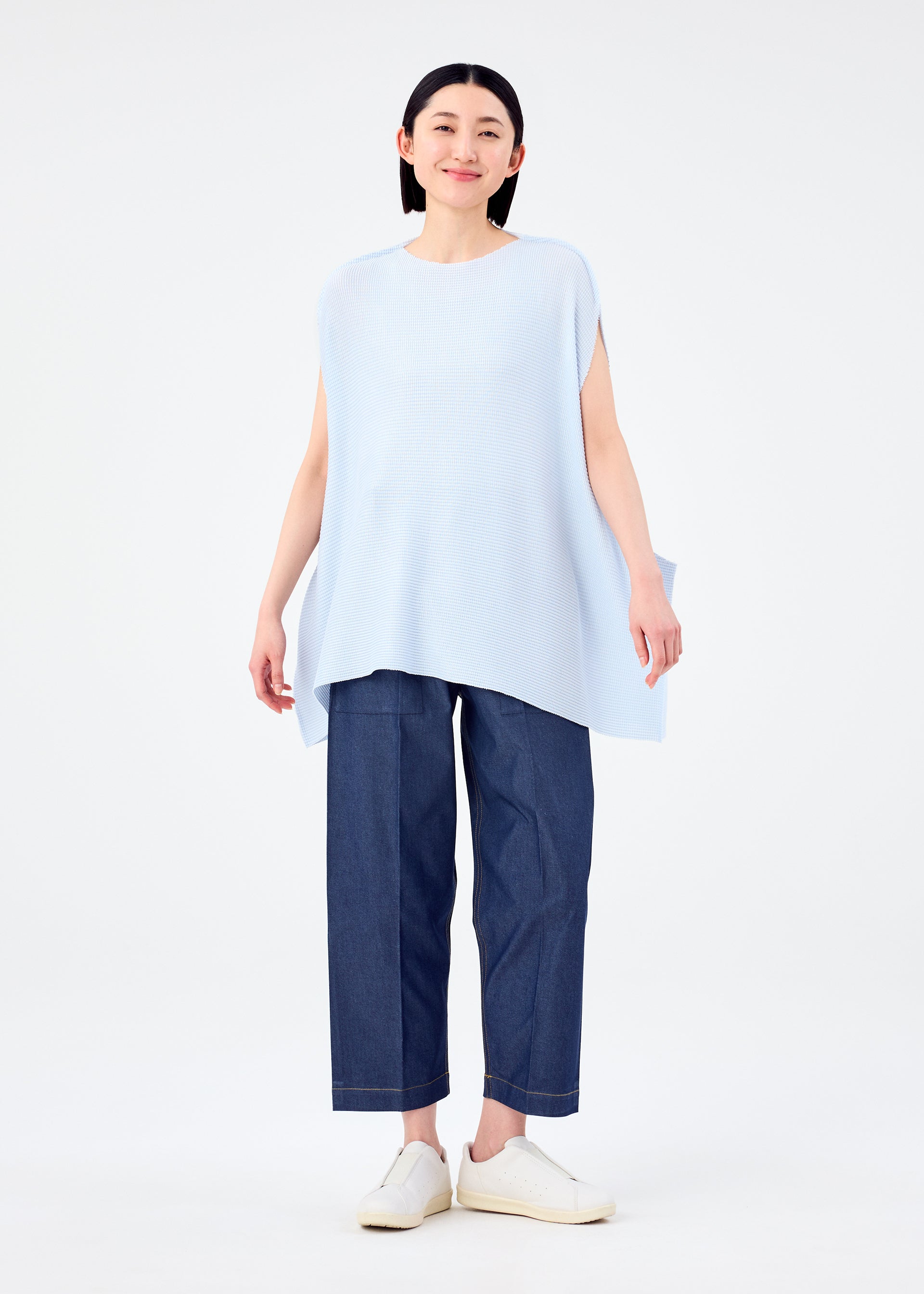Zoomed view A person with straight dark hair is smiling and wearing the me ISSEY MIYAKE CURVE KNIT STRETCH PLEATS top in light blue, paired with wide-leg dark blue jeans and white slip-on shoes, standing against a plain white background.