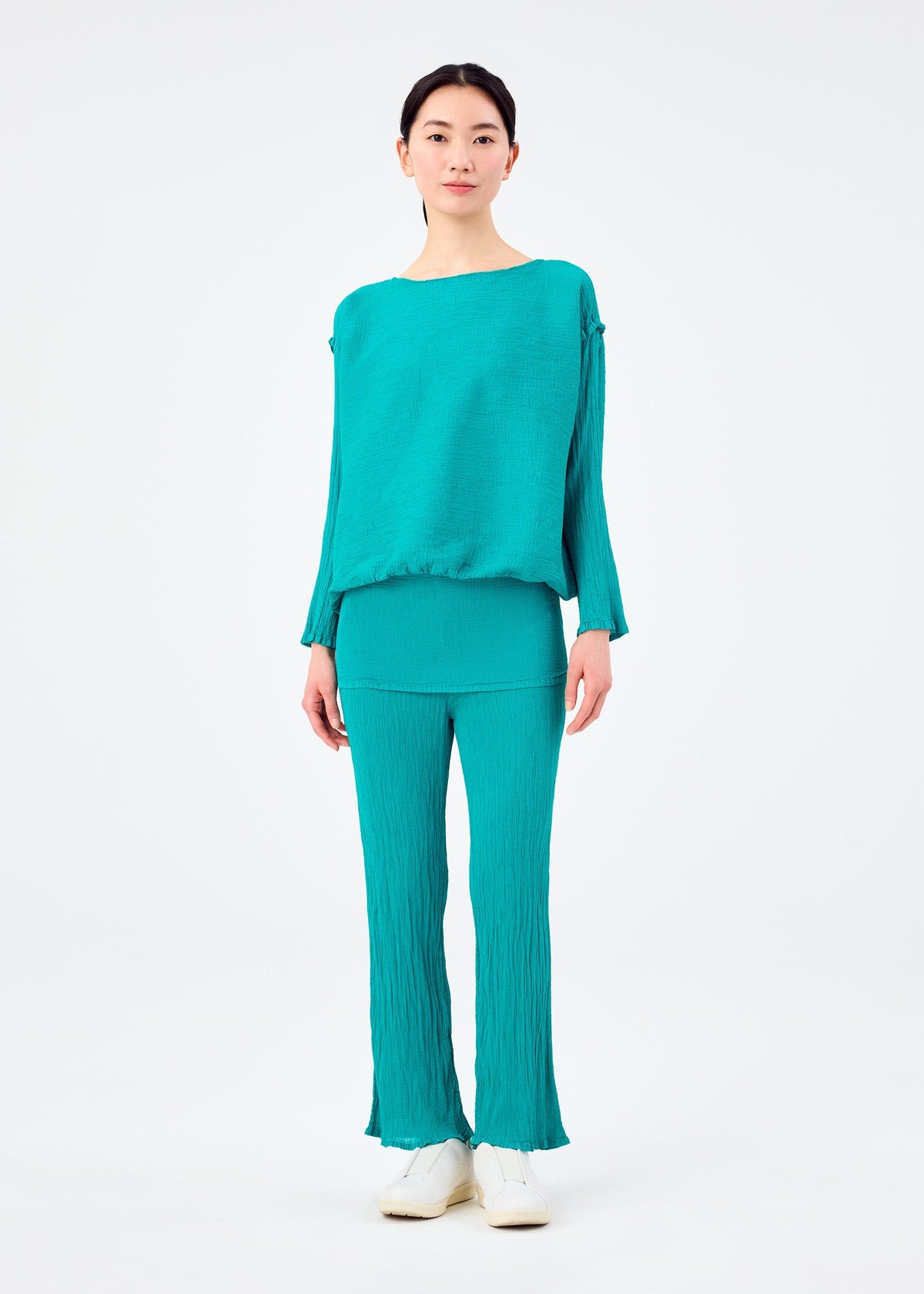 A person stands facing forward in the BALLOON SEE-THROUGH CREPE set by me ISSEY MIYAKE—a loose-fitting, teal top and pants made from crinkled high-twist thread. They wear white shoes and have short dark hair, against a plain white background. view 5 of 6