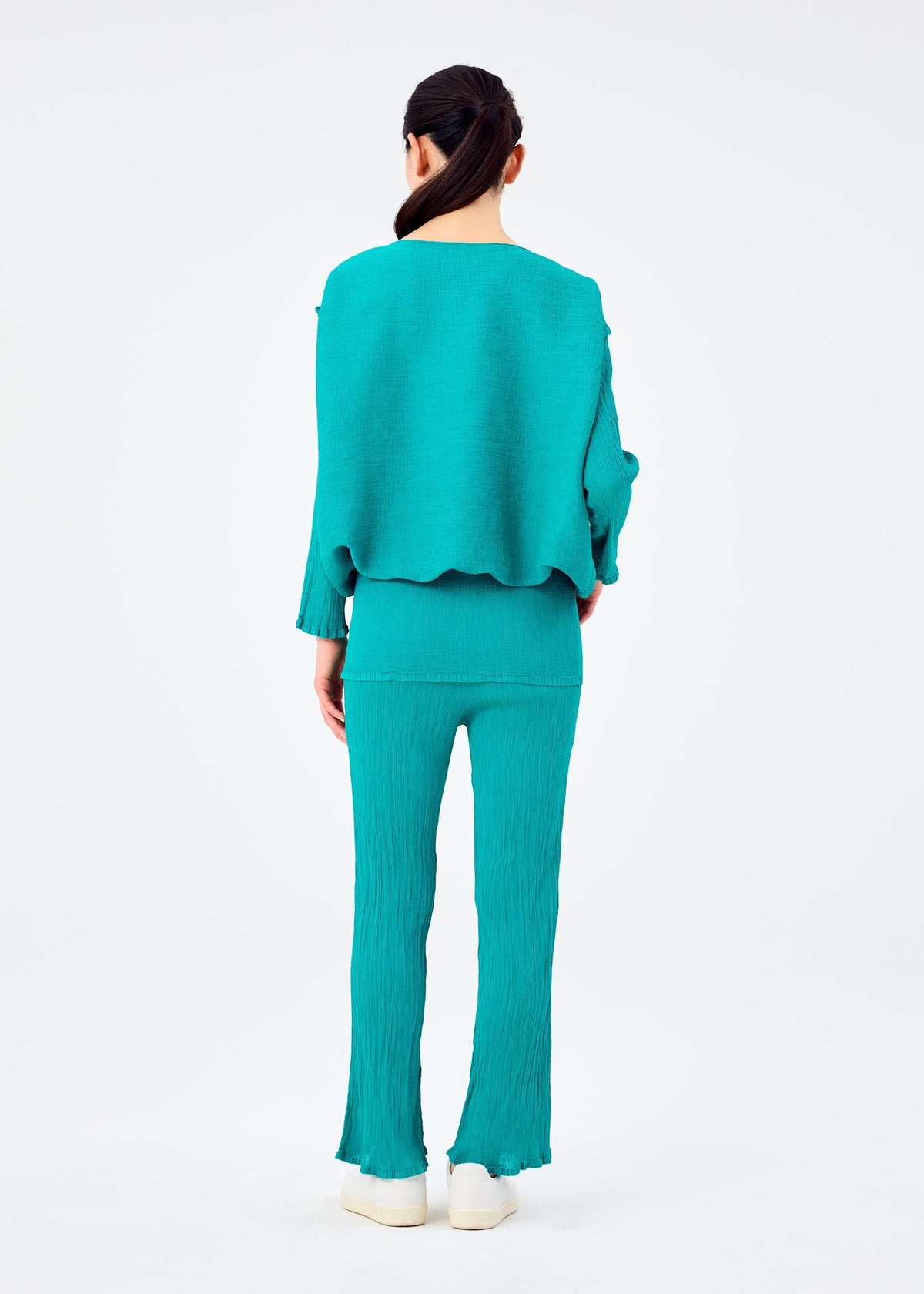 A person with long, dark hair tied back wears a loose top and turquoise me ISSEY MIYAKE BALLOON SEE-THROUGH CREPE pants with flared hems, plus white shoes, shown from behind against a plain white background. view 6 of 6