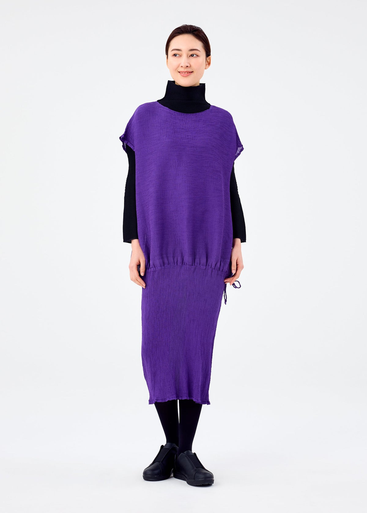 A woman smiles in the me ISSEY MIYAKE BALLOON SEE-THROUGH CREPE, a loose, purple short-sleeved dress worn over a black turtleneck and tights with black shoes, standing against a plain white background. view 5 of 6