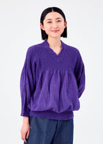 A woman with straight black hair wears the loose-fitting BALLOON CAULIFLOWER long-sleeved top by me ISSEY MIYAKE, paired with blue pants, standing against a white background and smiling gently at the camera.