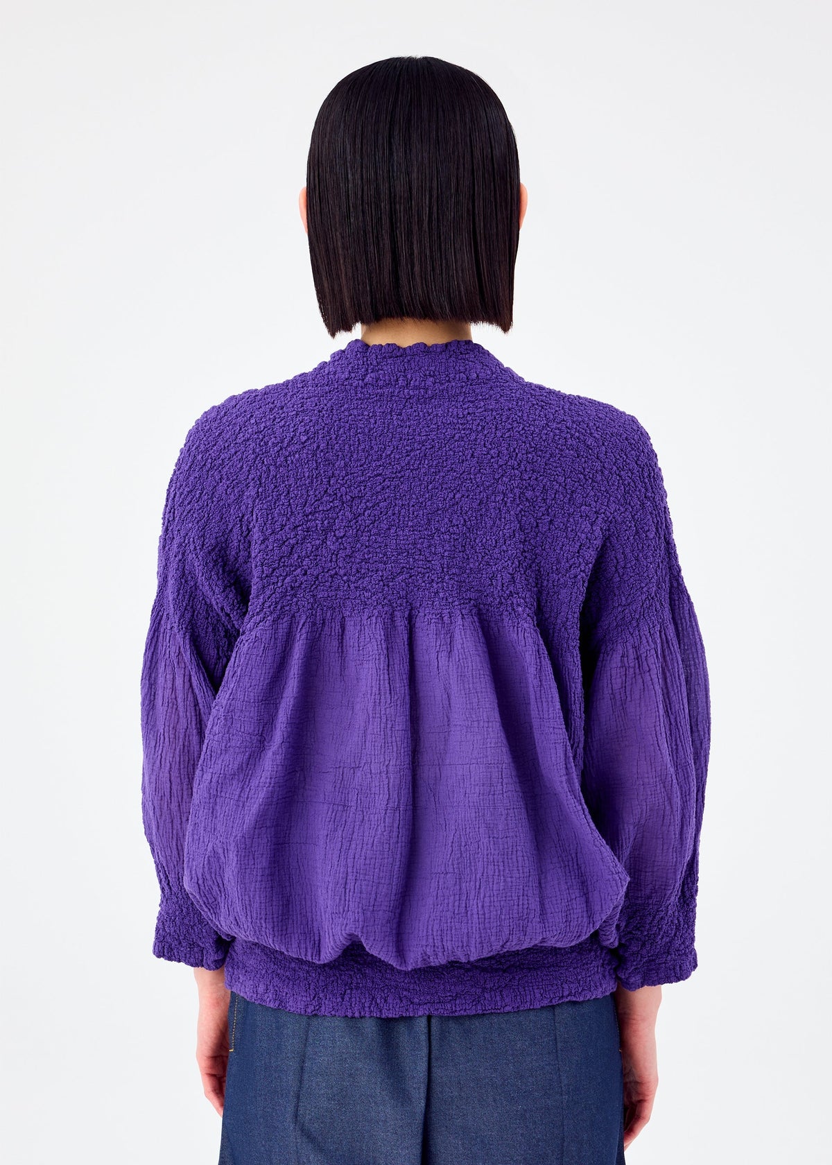 A person with straight, dark hair is seen from the back wearing the me ISSEY MIYAKE BALLOON CAULIFLOWER—a loose-fitting, purple blouse with a knit-like texture and gathered details—against a plain white background. view 6 of 7