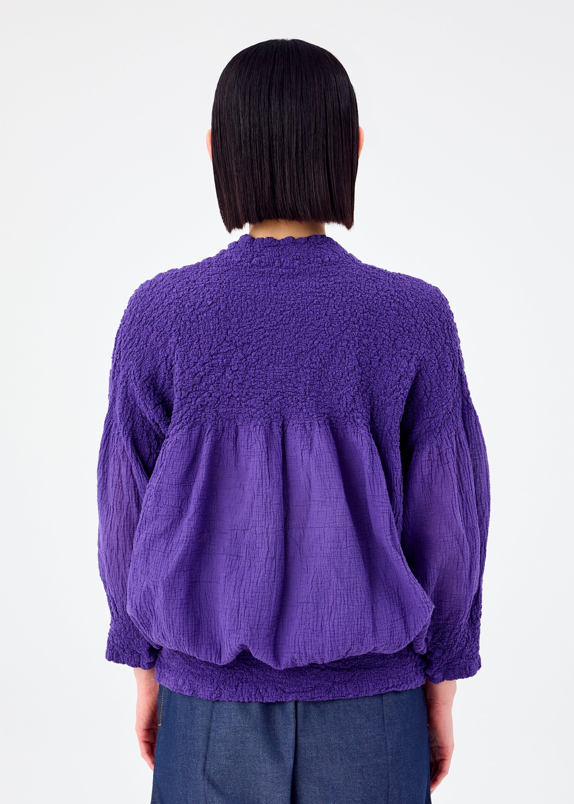 Zoomed view A person with straight, dark hair is seen from the back wearing the me ISSEY MIYAKE BALLOON CAULIFLOWER—a loose-fitting, purple blouse with a knit-like texture and gathered details—against a plain white background.
