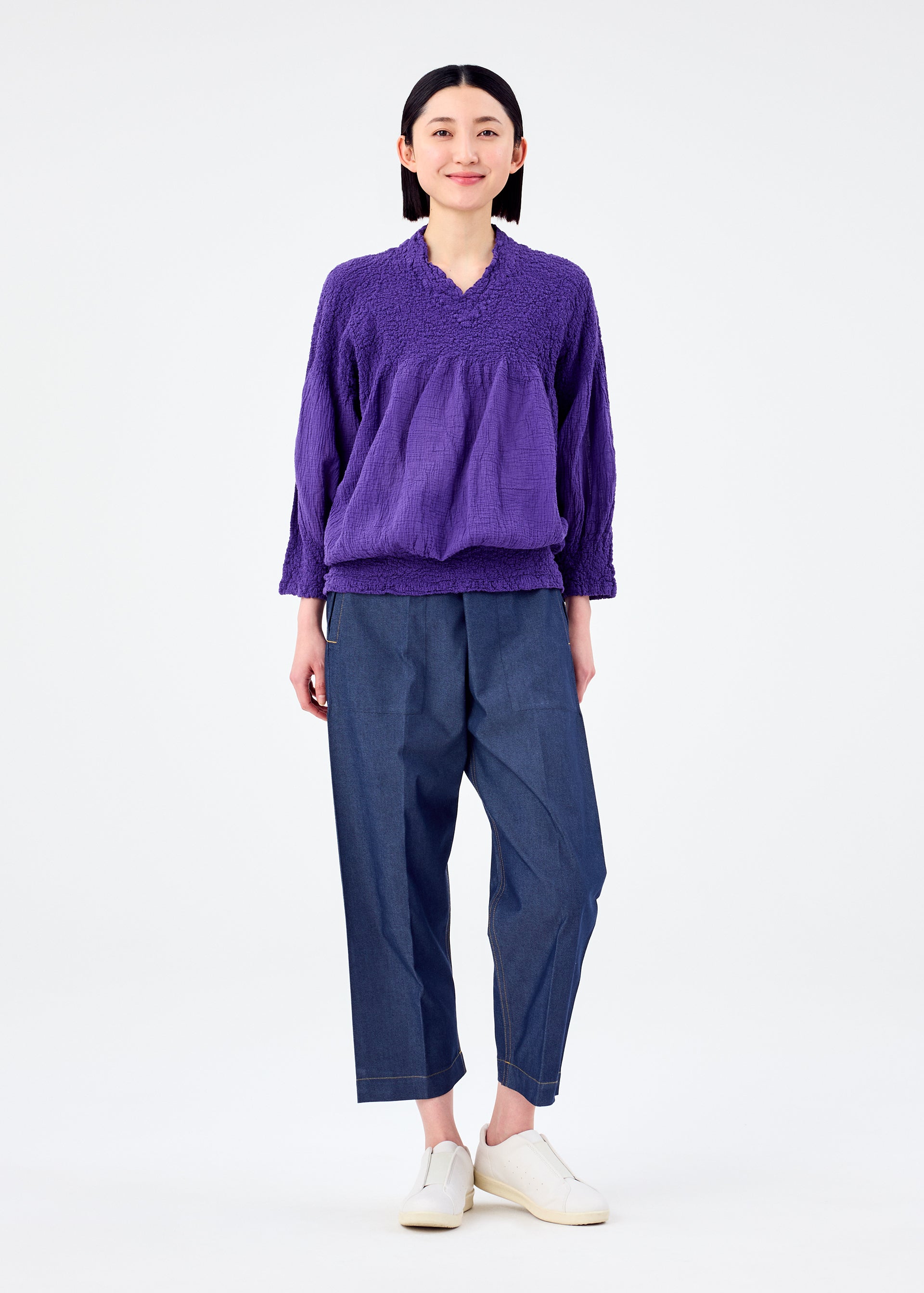 Zoomed view A person with short dark hair smiles against a white background, wearing the me ISSEY MIYAKE BALLOON CAULIFLOWER long-sleeved top, blue wide-leg pants, and white slip-on shoes.