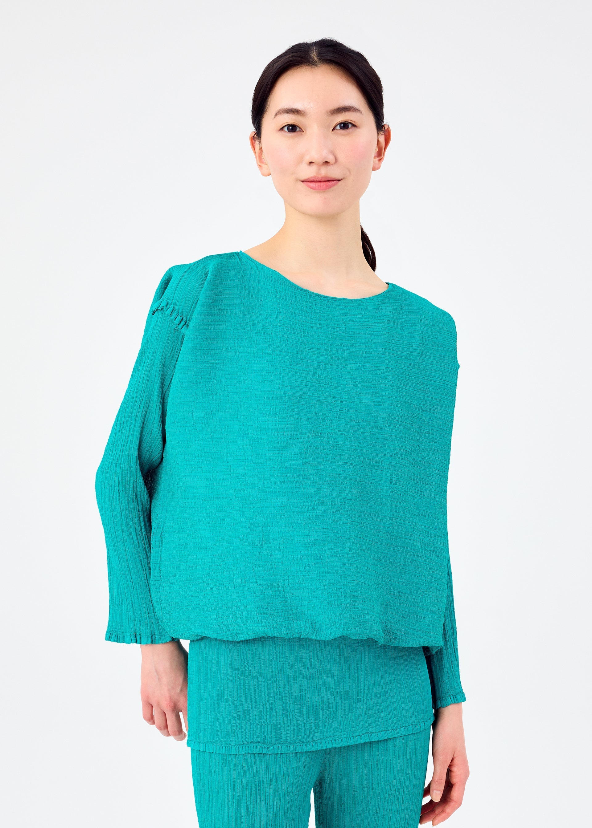 Zoomed view A person with long dark hair tied back wears the me ISSEY MIYAKE BALLOON SEE-THROUGH CREPE: a loose-fitting, long-sleeved turquoise blouse with matching pants, standing against a plain white background.