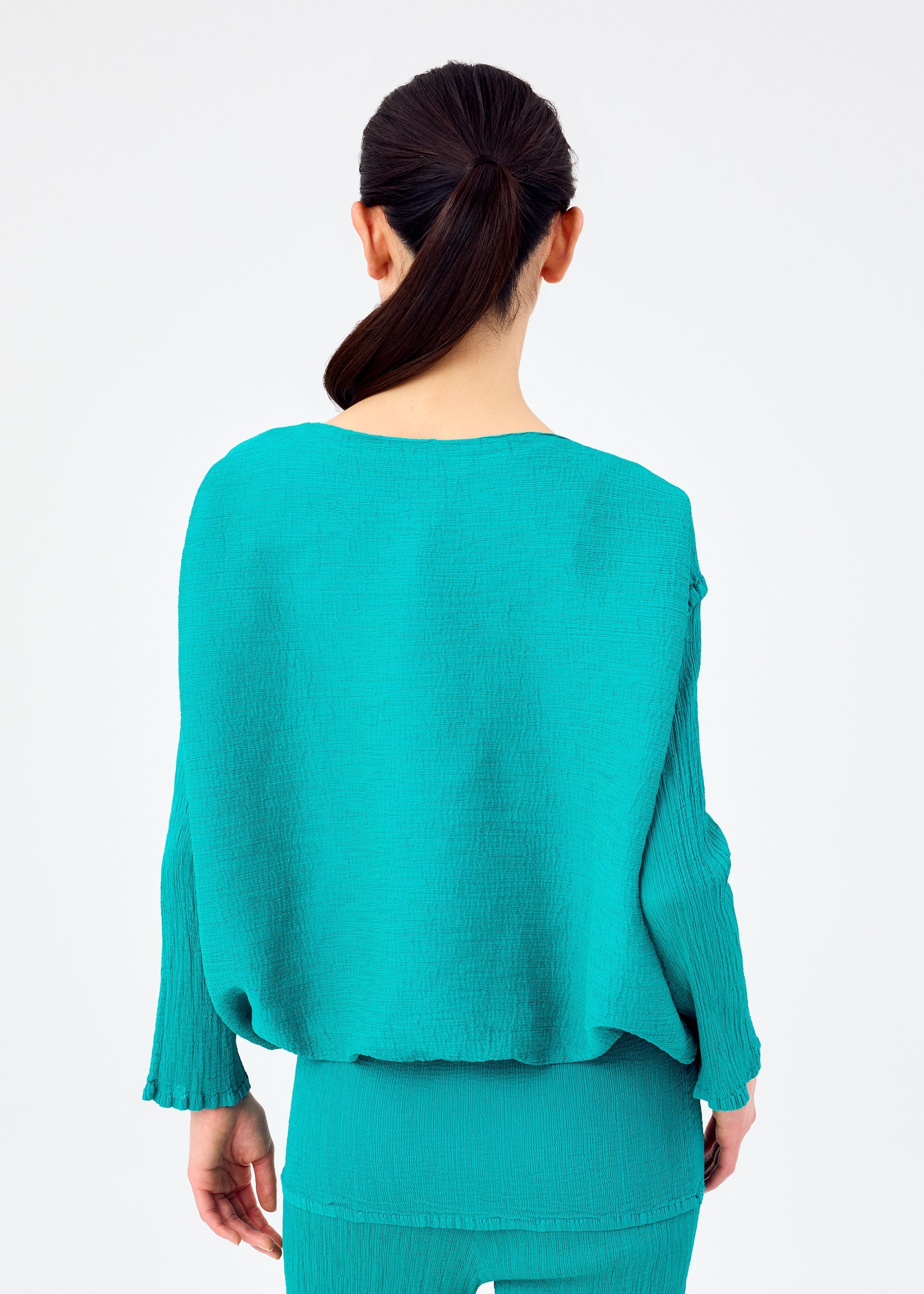 Zoomed view A woman with dark hair tied back is shown from behind, wearing the me ISSEY MIYAKE BALLOON SEE-THROUGH CREPE set in turquoise against a plain light background.