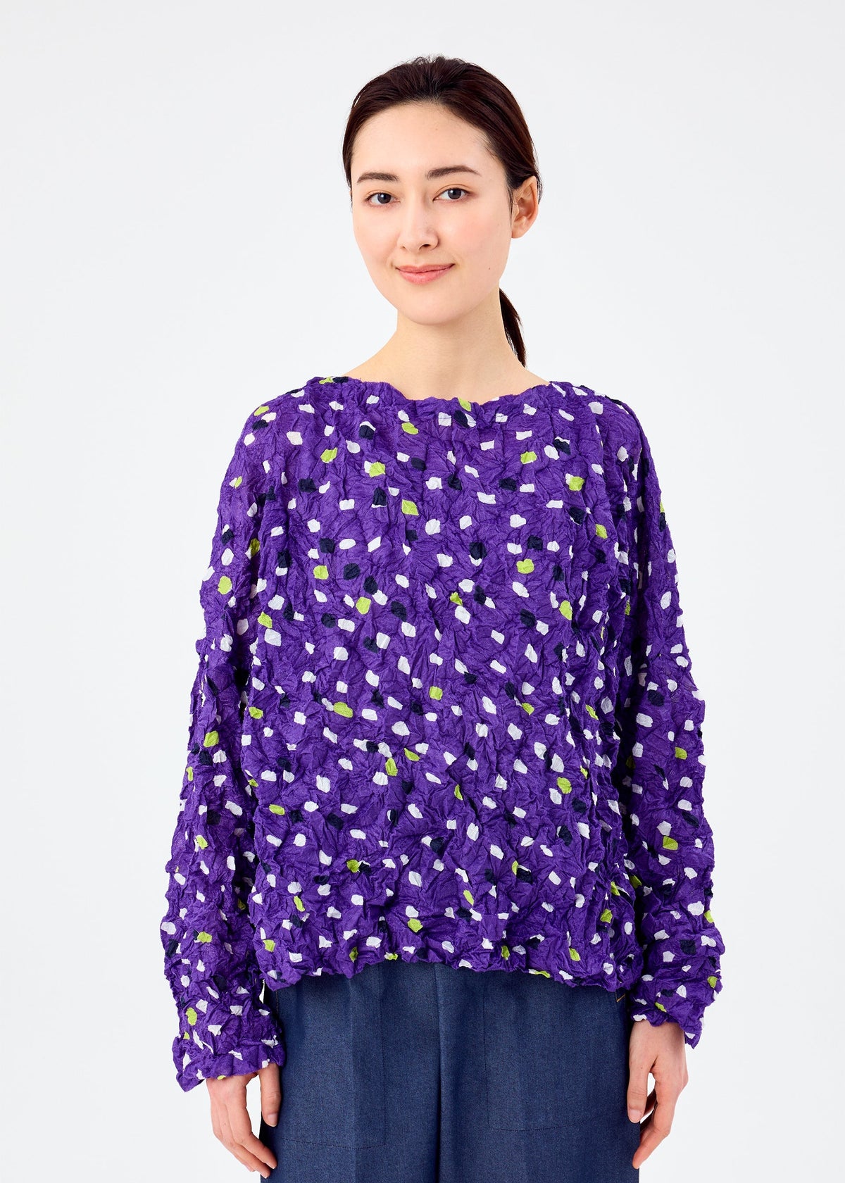 A woman with dark hair tied back smiles softly, wearing the TIME DOTS VELOUR MERINGUE top by me ISSEY MIYAKE with a white and green dotted pattern and dark blue pants, against a plain white background. view 5 of 7