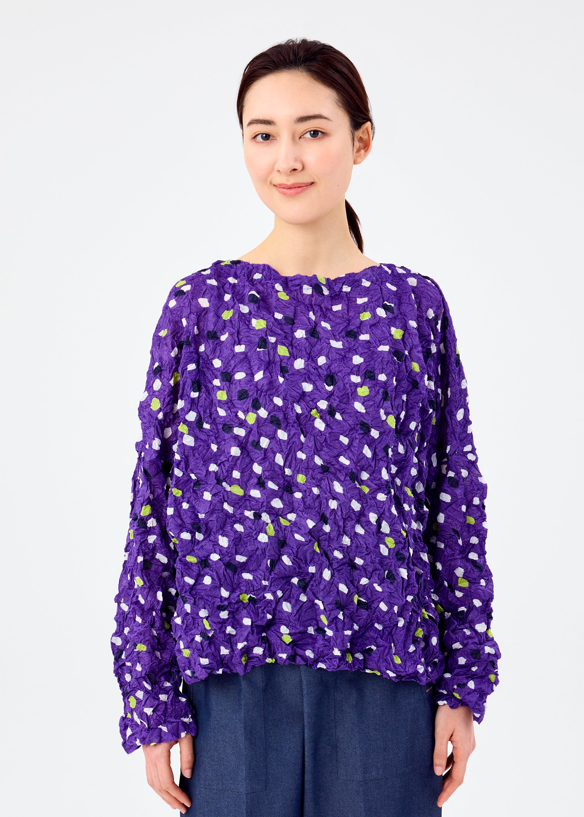 Zoomed view A woman with dark hair tied back smiles softly, wearing the TIME DOTS VELOUR MERINGUE top by me ISSEY MIYAKE with a white and green dotted pattern and dark blue pants, against a plain white background.