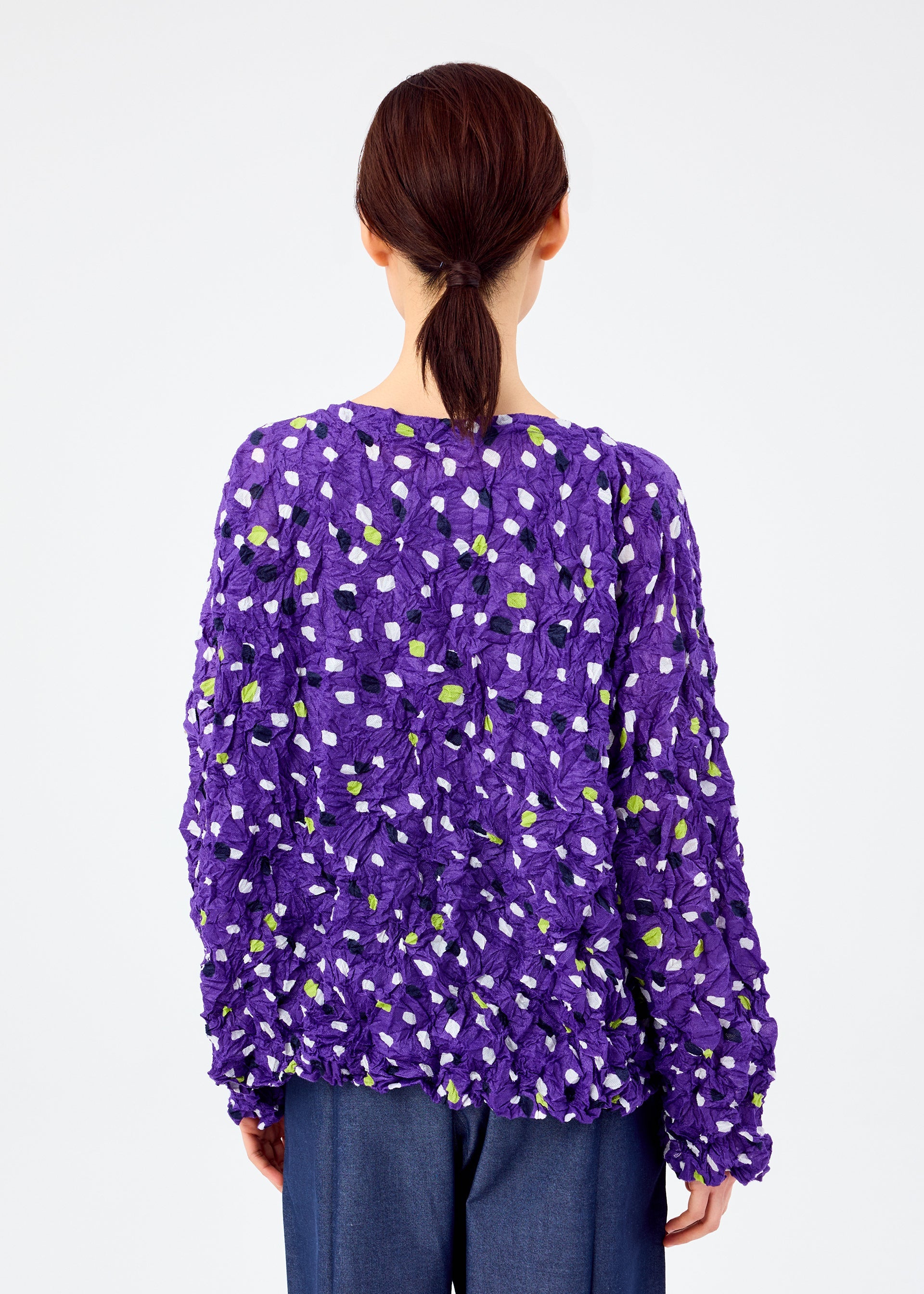 Zoomed view A person with brown hair in a low ponytail faces away, wearing the me ISSEY MIYAKE TIME DOTS VELOUR MERINGUE top—purple, green, and white dotted pattern—with dark blue pants on a plain white background.