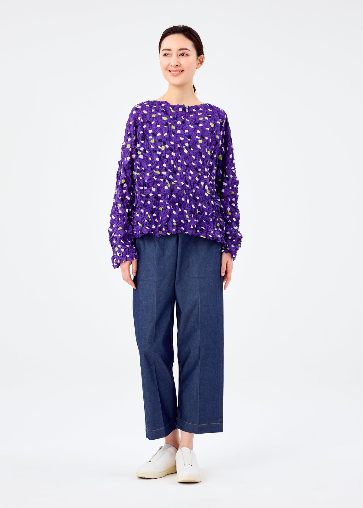 A woman wears the me ISSEY MIYAKE TIME DOTS VELOUR MERINGUE top against a white background, pairing it with wide-leg blue pants and white slip-on shoes. She smiles and gazes slightly to the side. view 7 of 7