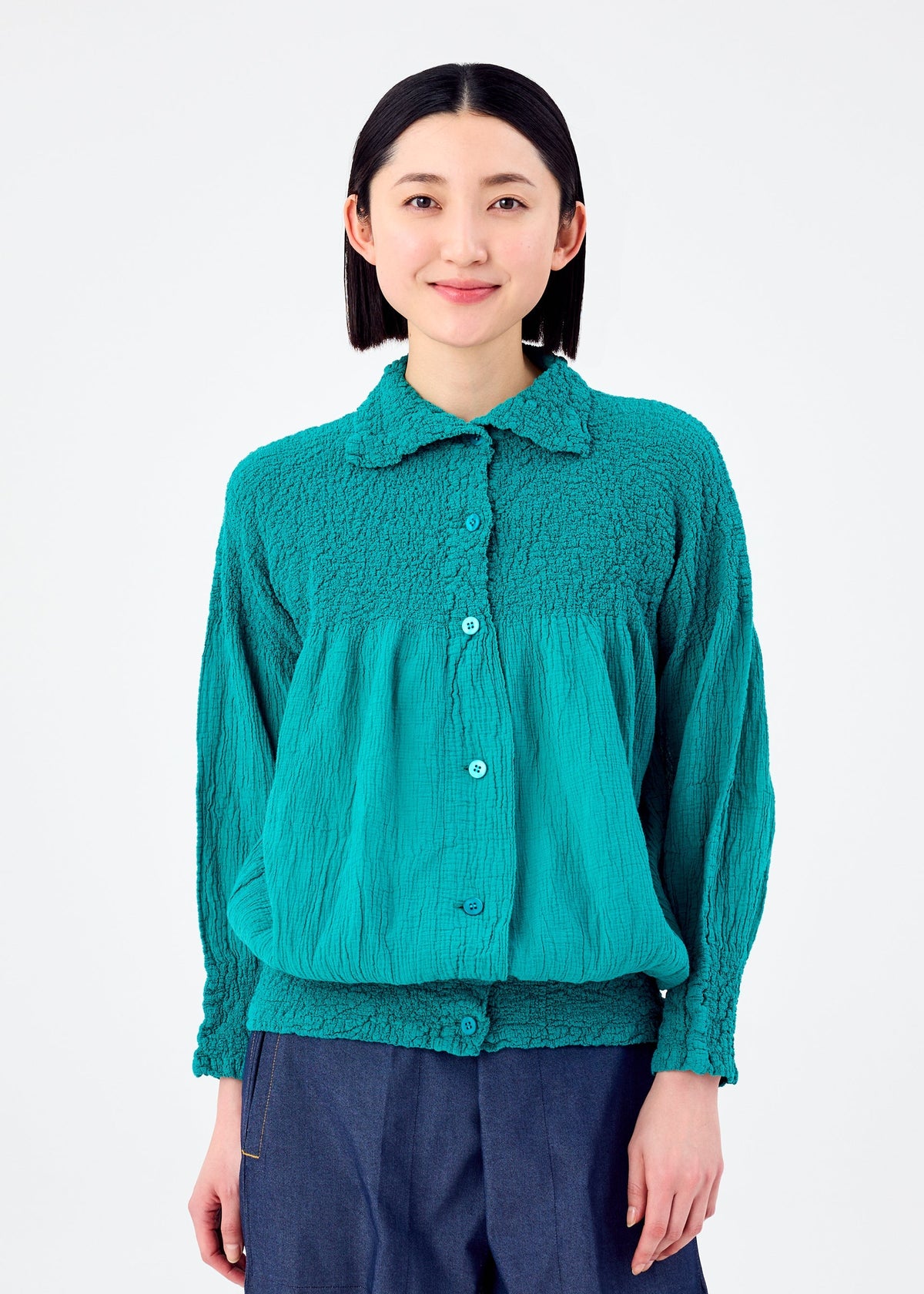 A person with shoulder-length black hair wears the BALLOON CAULIFLOWER teal cotton cardigan by me ISSEY MIYAKE and loose dark pants, standing against a plain white background. view 5 of 7