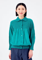A person with shoulder-length black hair wears the BALLOON CAULIFLOWER teal cotton cardigan by me ISSEY MIYAKE and loose dark pants, standing against a plain white background.