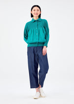 A person stands smiling with hands behind their back, wearing the BALLOON CAULIFLOWER cardigan in teal by me ISSEY MIYAKE, wide-leg dark blue pants, and white slip-on shoes against a white background.