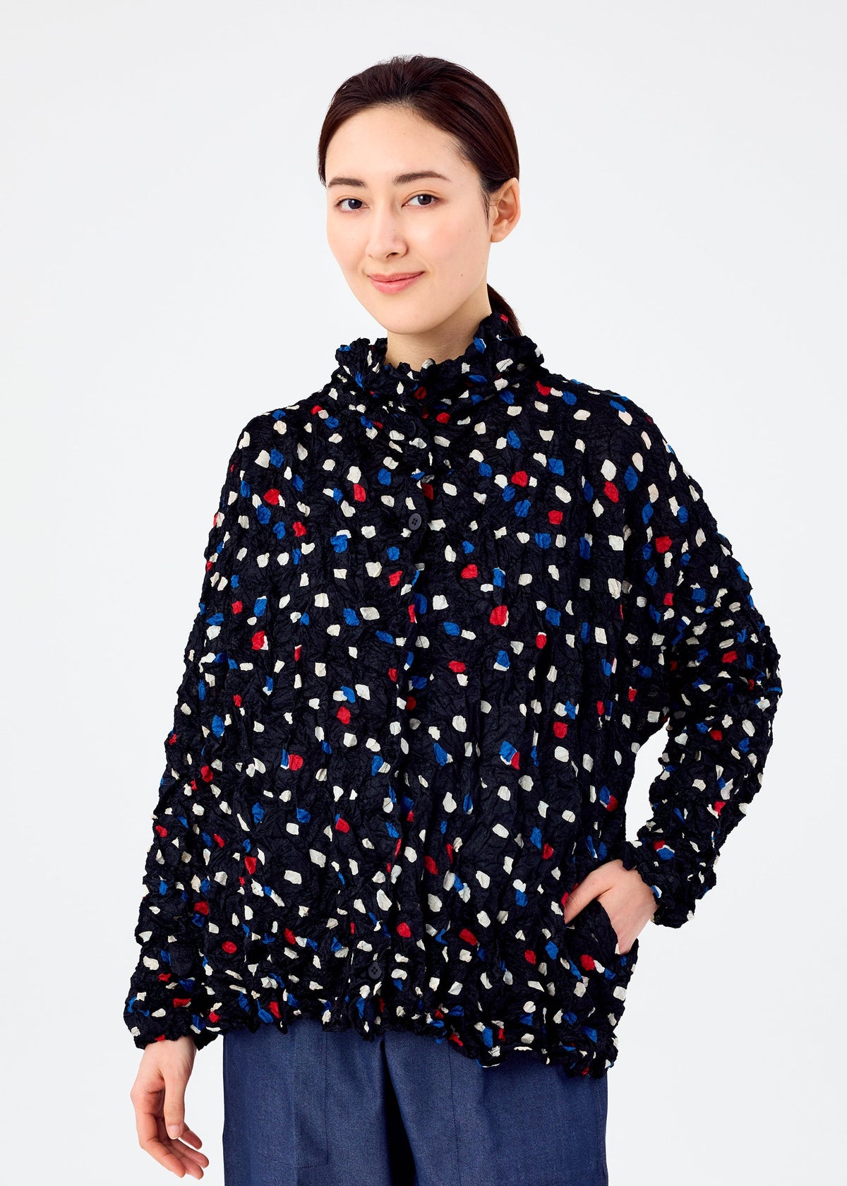A person with dark hair pulled back wears the me ISSEY MIYAKE TIME DOTS VELOUR MERINGUE cardigan, featuring a colorful dotted pattern, paired with blue pants, standing against a plain white background and smiling slightly. view 6 of 8