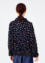 Shown from behind, a person with brown hair tied back wears the TIME DOTS VELOUR MERINGUE cardigan by me ISSEY MIYAKE, featuring red, blue, and white dots, paired with blue pants against a plain light background.