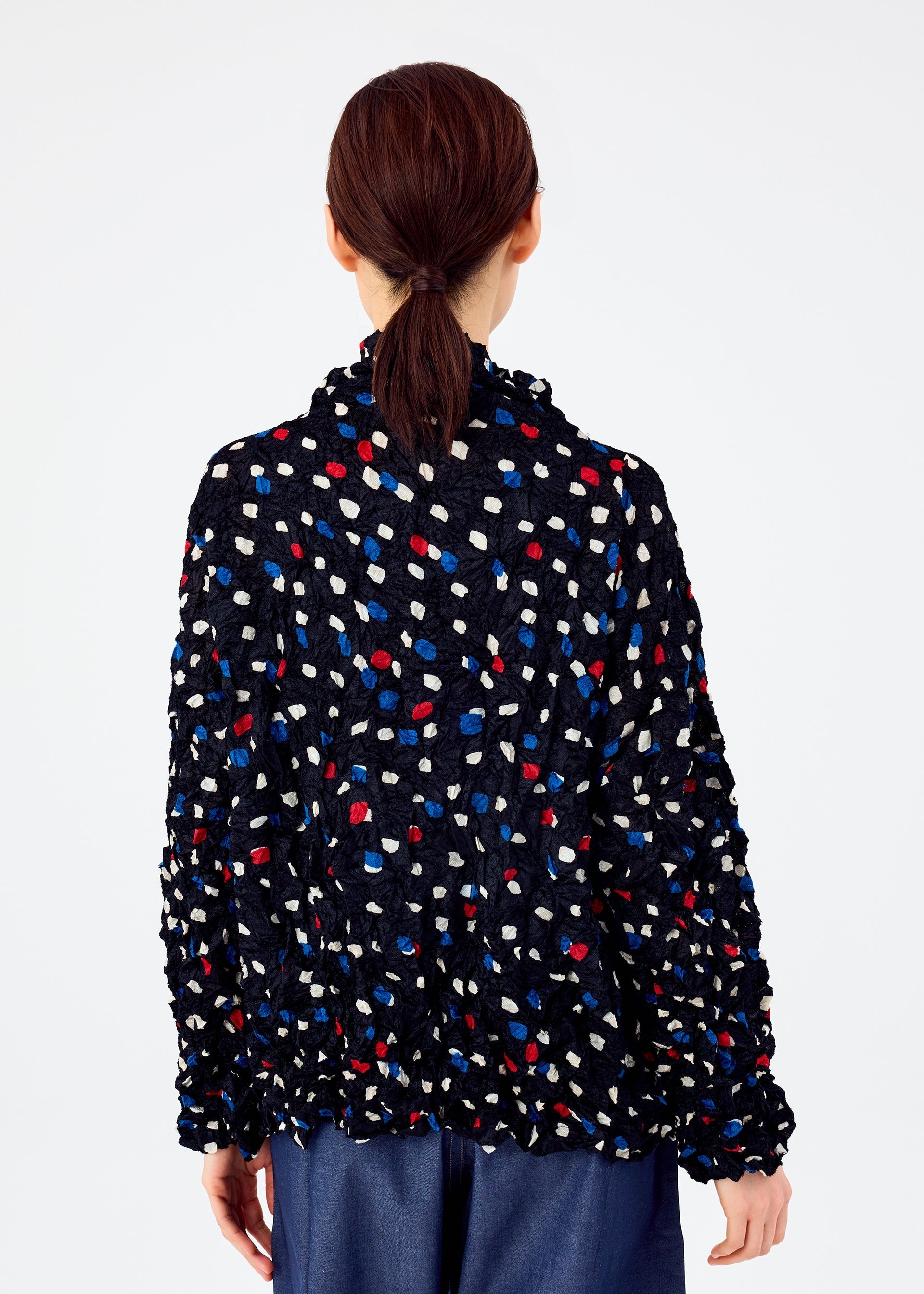 Zoomed view Shown from behind, a person with brown hair tied back wears the TIME DOTS VELOUR MERINGUE cardigan by me ISSEY MIYAKE, featuring red, blue, and white dots, paired with blue pants against a plain light background.