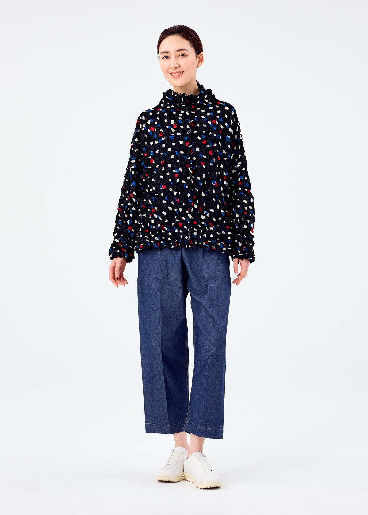 A person stands smiling in the TIME DOTS VELOUR MERINGUE cardigan by me ISSEY MIYAKE, featuring a dotted pattern, paired with loose blue jeans and white shoes against a plain white background. view 8 of 8
