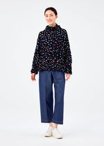 A person stands smiling in the TIME DOTS VELOUR MERINGUE cardigan by me ISSEY MIYAKE, featuring a dotted pattern, paired with loose blue jeans and white shoes against a plain white background.