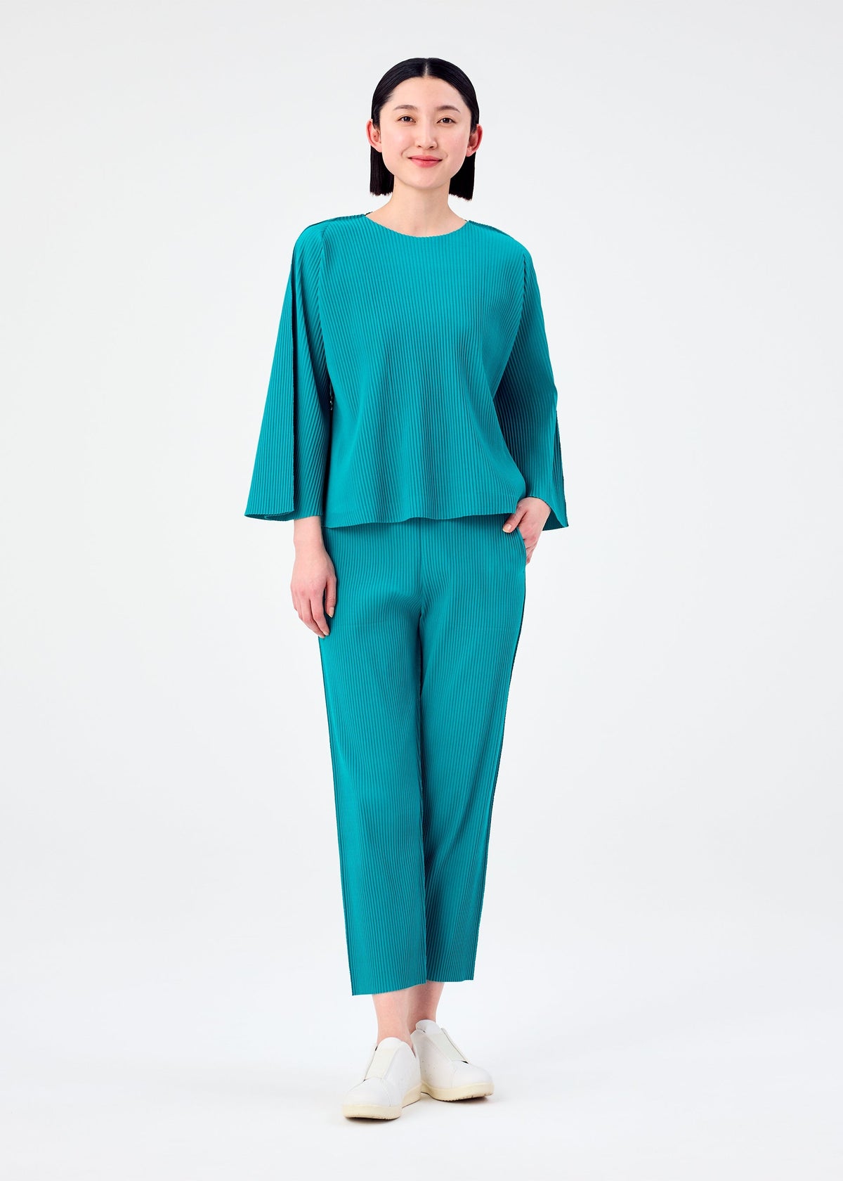 A person stands against a plain white background, wearing the me ISSEY MIYAKE SWING PLEATS—a loose-fitting teal top with long sleeves and lightweight straight pants—paired with white slip-on shoes, smiling softly. view 4 of 5