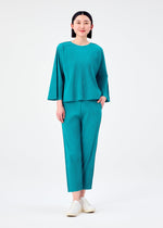 A person stands against a plain white background, wearing the me ISSEY MIYAKE SWING PLEATS—a loose-fitting teal top with long sleeves and lightweight straight pants—paired with white slip-on shoes, smiling softly.