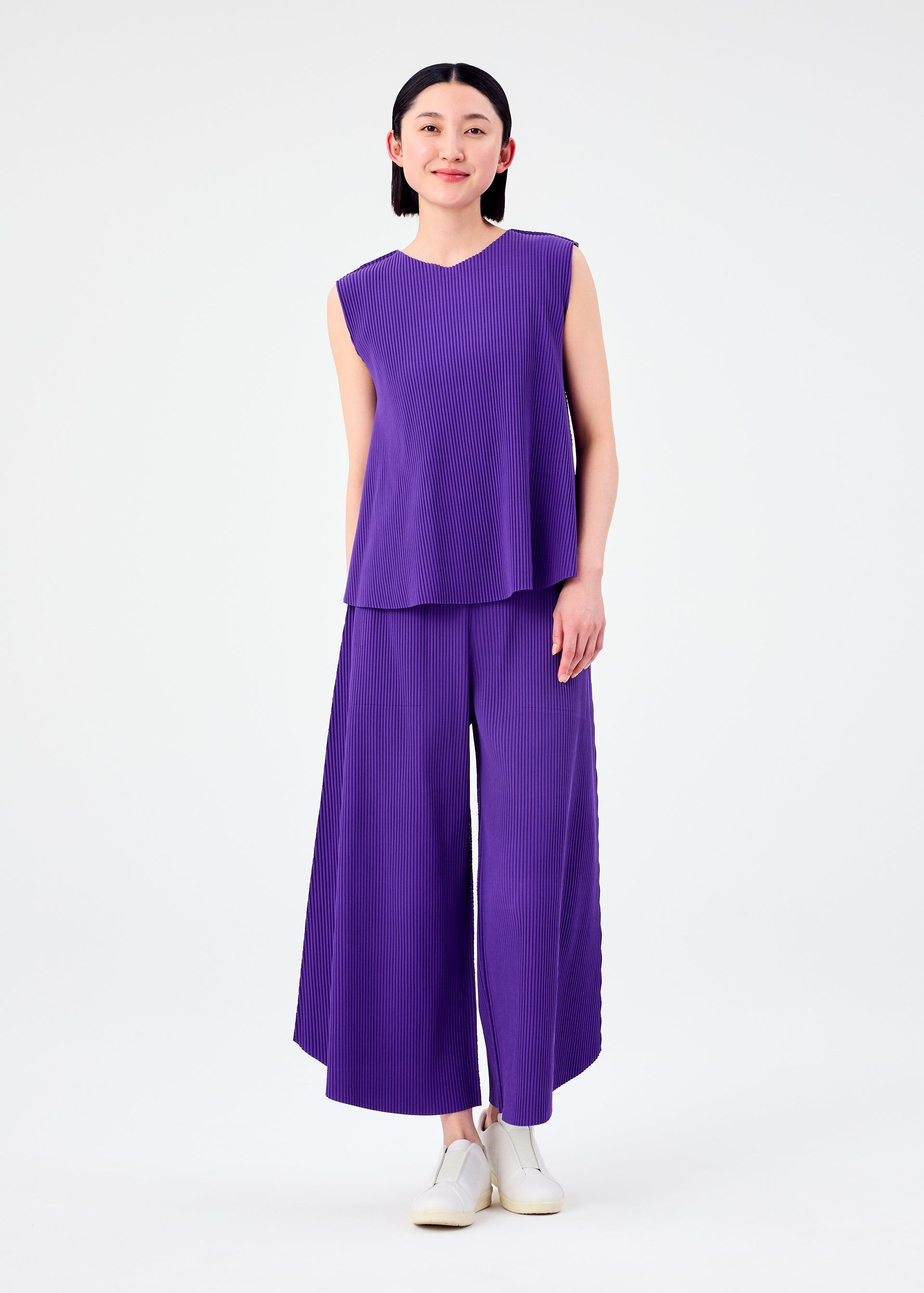 Zoomed view A person smiles against a white background wearing the SWING PLEATS sleeveless, loose-fit purple top and matching pleated knit pants by me ISSEY MIYAKE, paired with white slip-on shoes. They have straight, dark hair.
