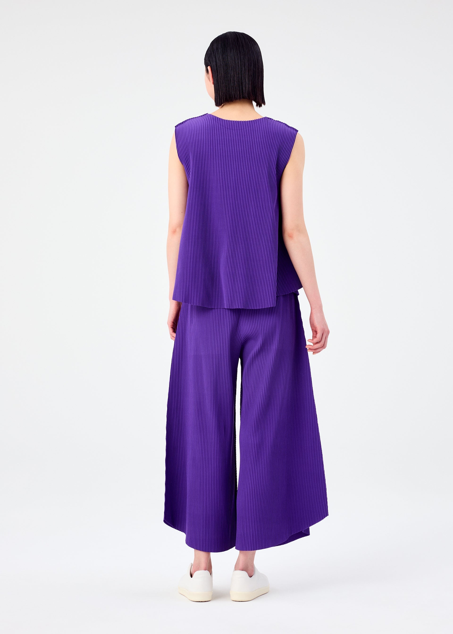 Zoomed view Seen from behind, a person with short dark hair wears the SWING PLEATS sleeveless top and matching pleated knit pants by me ISSEY MIYAKE in purple with fine pleats, white slip-on shoes, and stands against a plain white background.