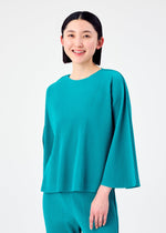 A woman with straight dark hair smiles while wearing the teal SWING PLEATS long-sleeved top and matching pleated pants by me ISSEY MIYAKE, standing against a plain white background.