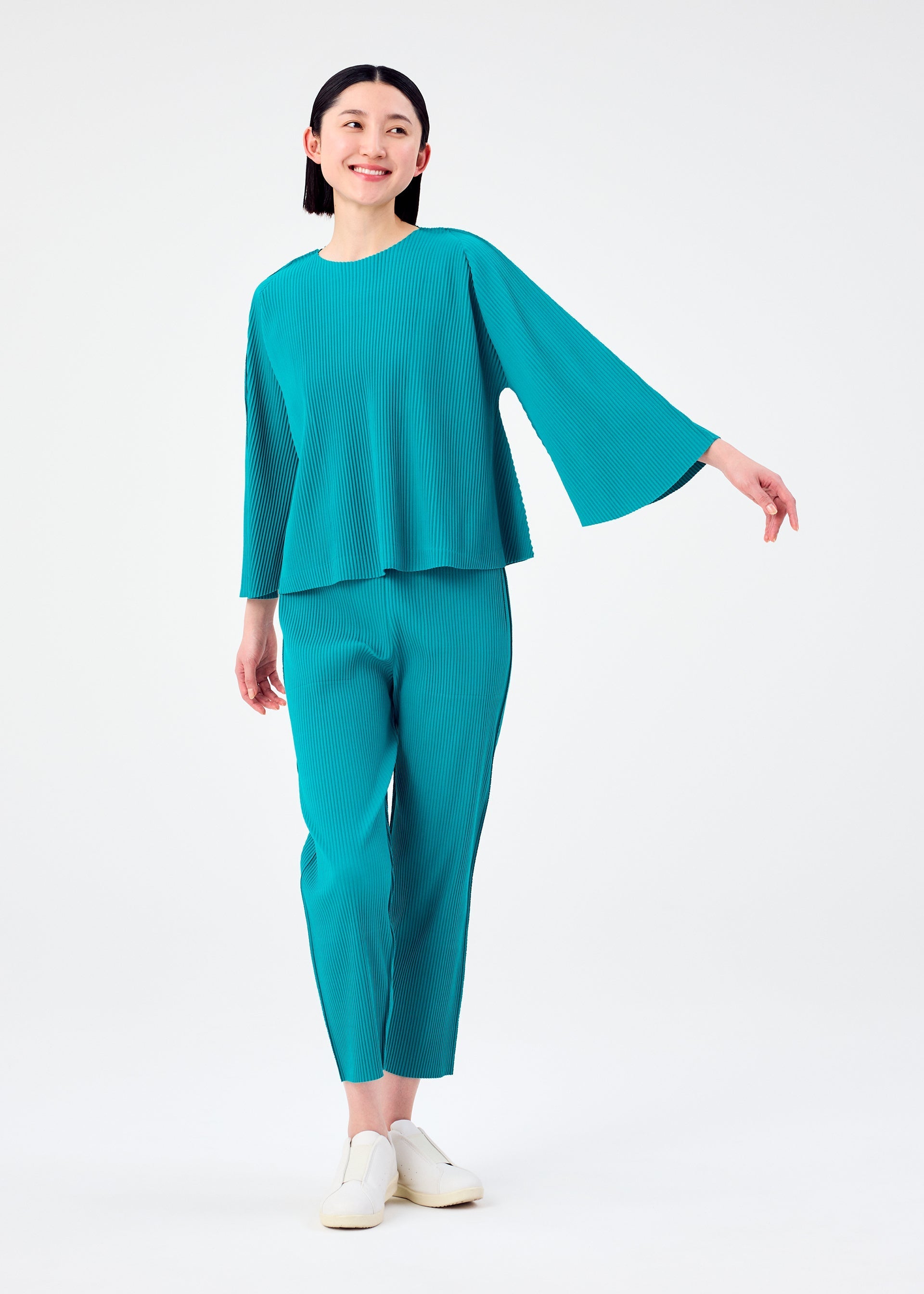 Zoomed view A woman smiles in the SWING PLEATS set by me ISSEY MIYAKE, featuring a teal, lightweight knit pleated loose-fit top with wide sleeves and matching tapered pants, styled with white slip-on shoes against a plain light background.