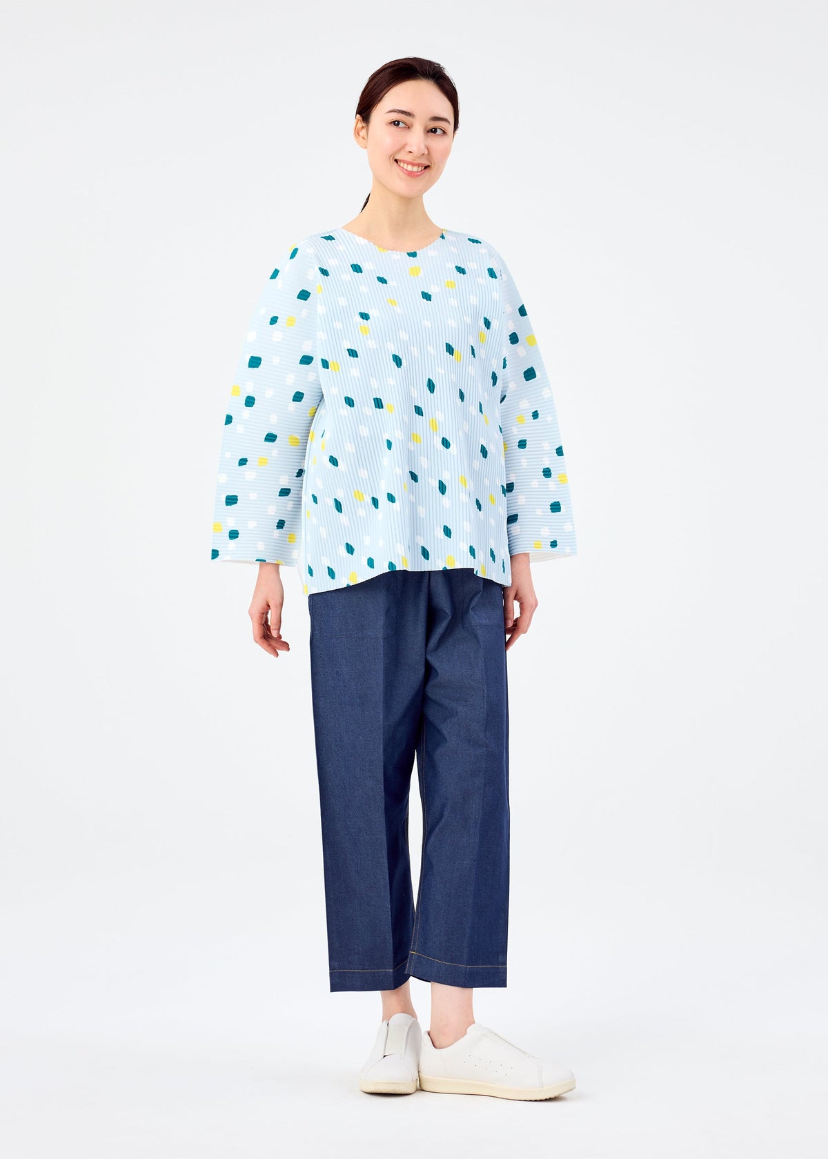 A woman smiles in the TIME DOTS FINE KNIT PLEATS T by me ISSEY MIYAKE—a light blue patterned long-sleeve top with colorful dots—paired with loose dark blue pants and white slip-on shoes, set against a plain white background. view 7 of 8