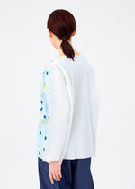 A person with straight brown hair in a low ponytail wears the me ISSEY MIYAKE TIME DOTS FINE KNIT PLEATS T, a loose, long-sleeved top with subtle blue and yellow accents, paired with dark pants against a plain light background.