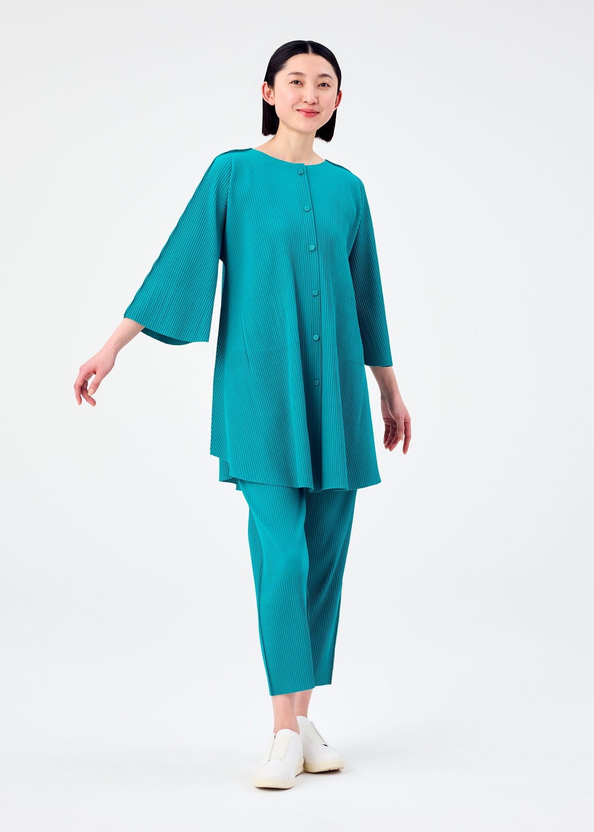 A woman smiles, wearing the SWING PLEATS loose turquoise tunic and matching pants by me ISSEY MIYAKE. She has straight dark hair and white slip-on shoes, posing against a plain white background. view 7 of 9