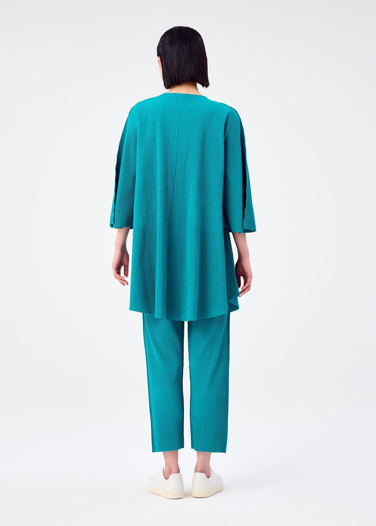 A person with straight, dark hair faces away, wearing the teal SWING PLEATS top and matching pants by me ISSEY MIYAKE, paired with white slip-on shoes against a plain white background. view 8 of 9