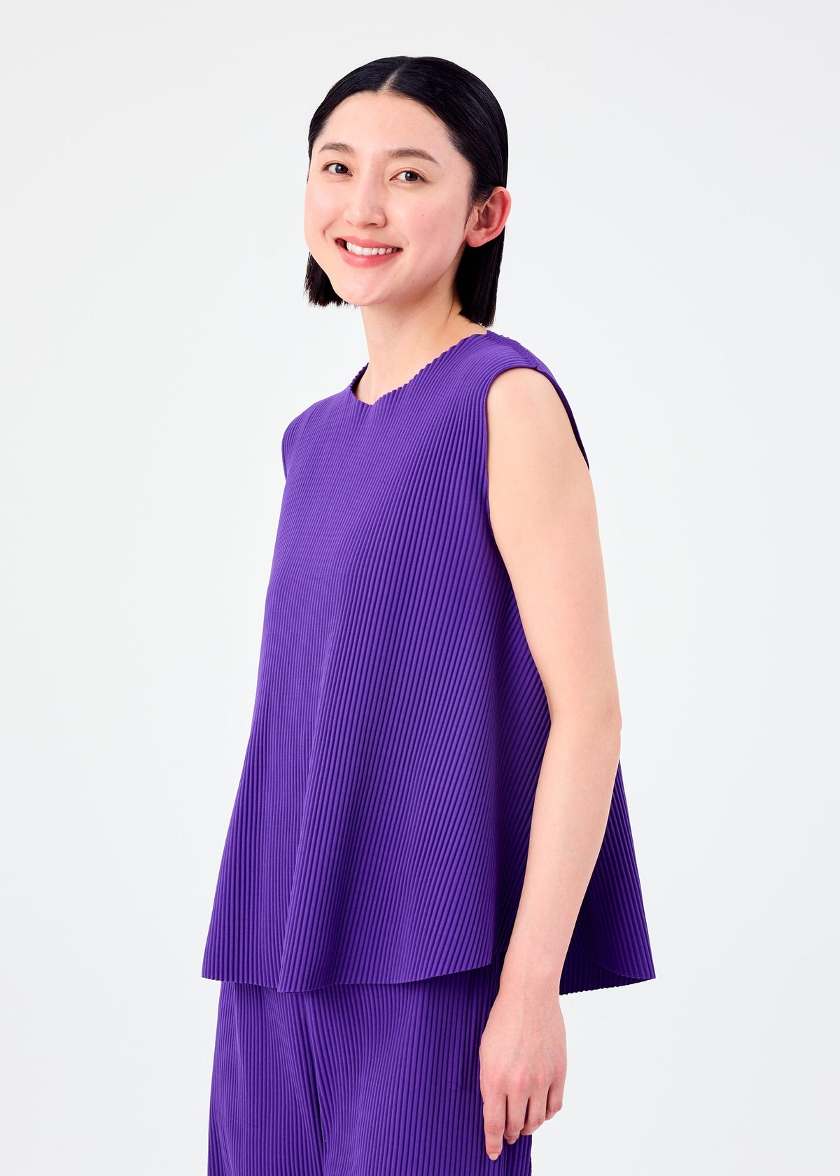 A woman with straight dark hair, wearing the SWING PLEATS sleeveless knit top and matching pants by me ISSEY MIYAKE, smiles at the camera against a plain white background. view 6 of 8