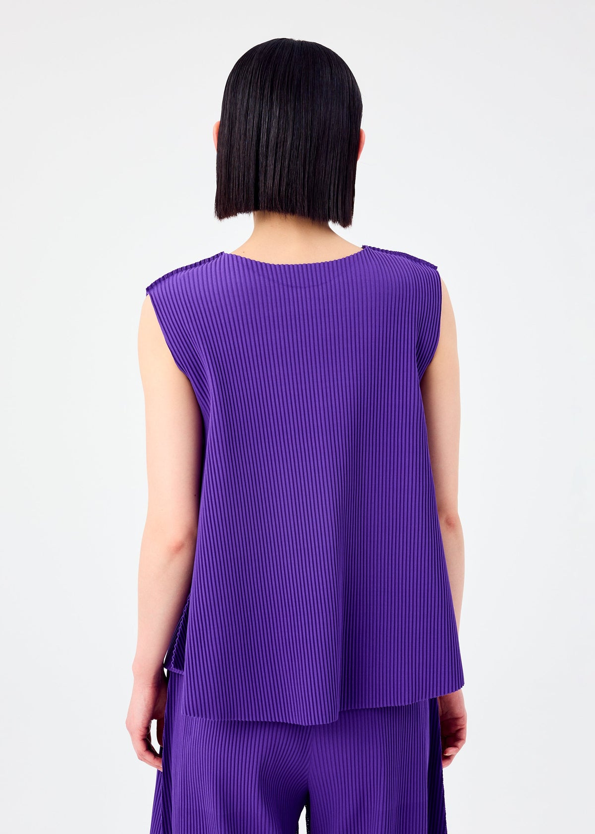 A person with straight, chin-length dark hair is shown from the back, wearing the me ISSEY MIYAKE SWING PLEATS sleeveless, loose-fitting pleated knit top and matching purple pants against a plain white background. view 7 of 8