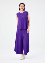 A person stands against a plain white background, wearing the SWING PLEATS sleeveless top and matching wide-leg pleated purple pants by me ISSEY MIYAKE, both featuring fine knit pleats, paired with white slip-on shoes.