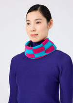 A woman with straight dark hair in a low bun wears the me ISSEY MIYAKE DIAMOND PLEATS STOLE, featuring bold pink and blue pleats. She smiles softly, facing slightly right against a plain light background.