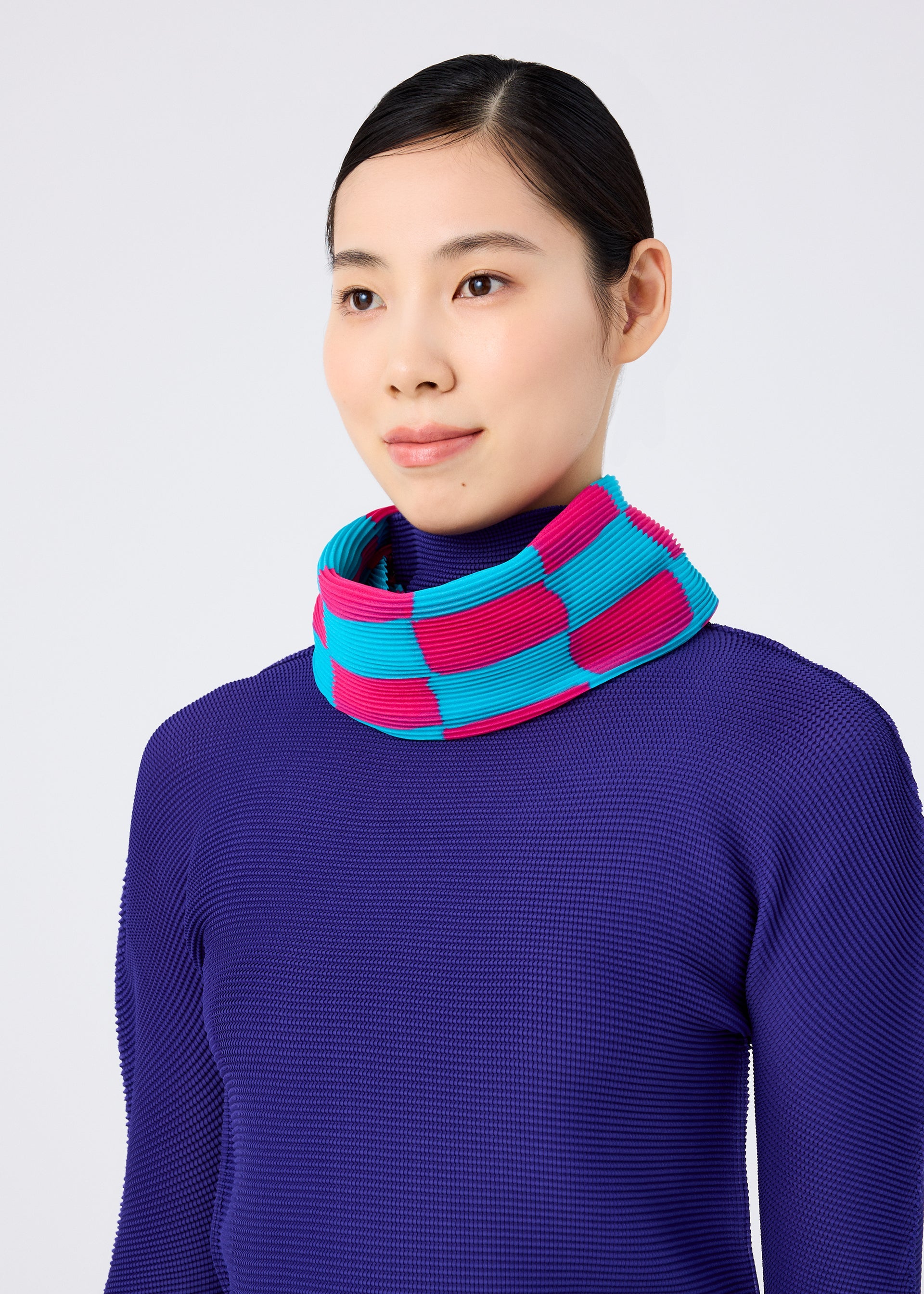 Zoomed view A woman with straight dark hair in a low bun wears the me ISSEY MIYAKE DIAMOND PLEATS STOLE, featuring bold pink and blue pleats. She smiles softly, facing slightly right against a plain light background.