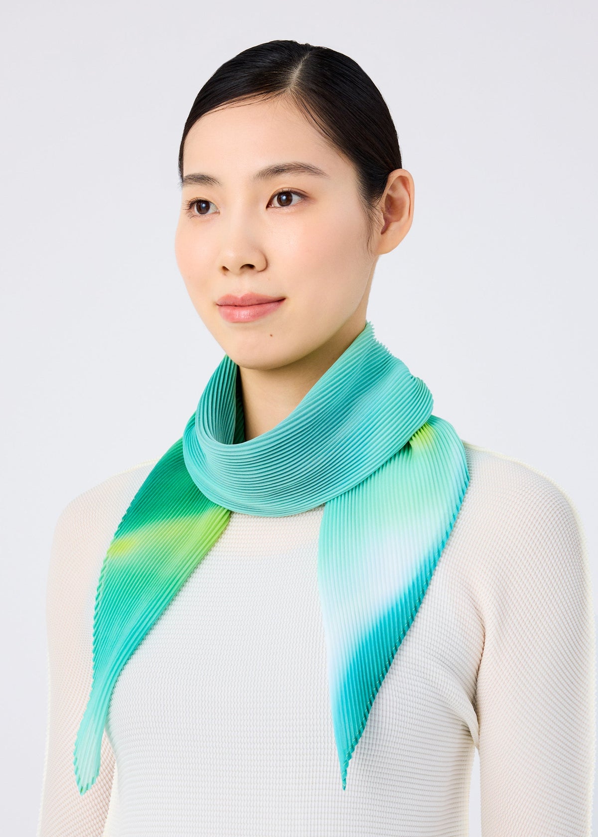 A woman with straight black hair pulled back stands against a plain light background, wearing a sheer white top and the me ISSEY MIYAKE AURORA PLEATS STOLE around her neck. view 5 of 5