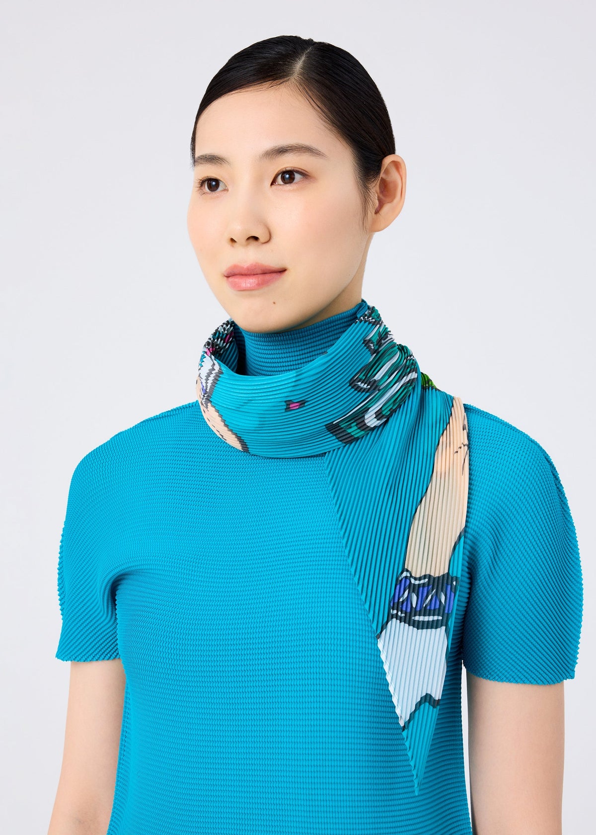 A woman with straight black hair in a neat bun wears a textured turquoise top and the MAGIC PLEATS STOLE by me ISSEY MIYAKE with a colorful print, wrapped around her neck. She gazes slightly to the side against a plain light background. view 5 of 5