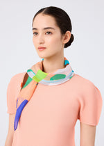 A woman with dark hair in a low bun wears a light pink textured top and the me ISSEY MIYAKE SURPRISE BOX PLEATS STOLE, showcasing its unique green, blue, and orange pattern against a plain light background.