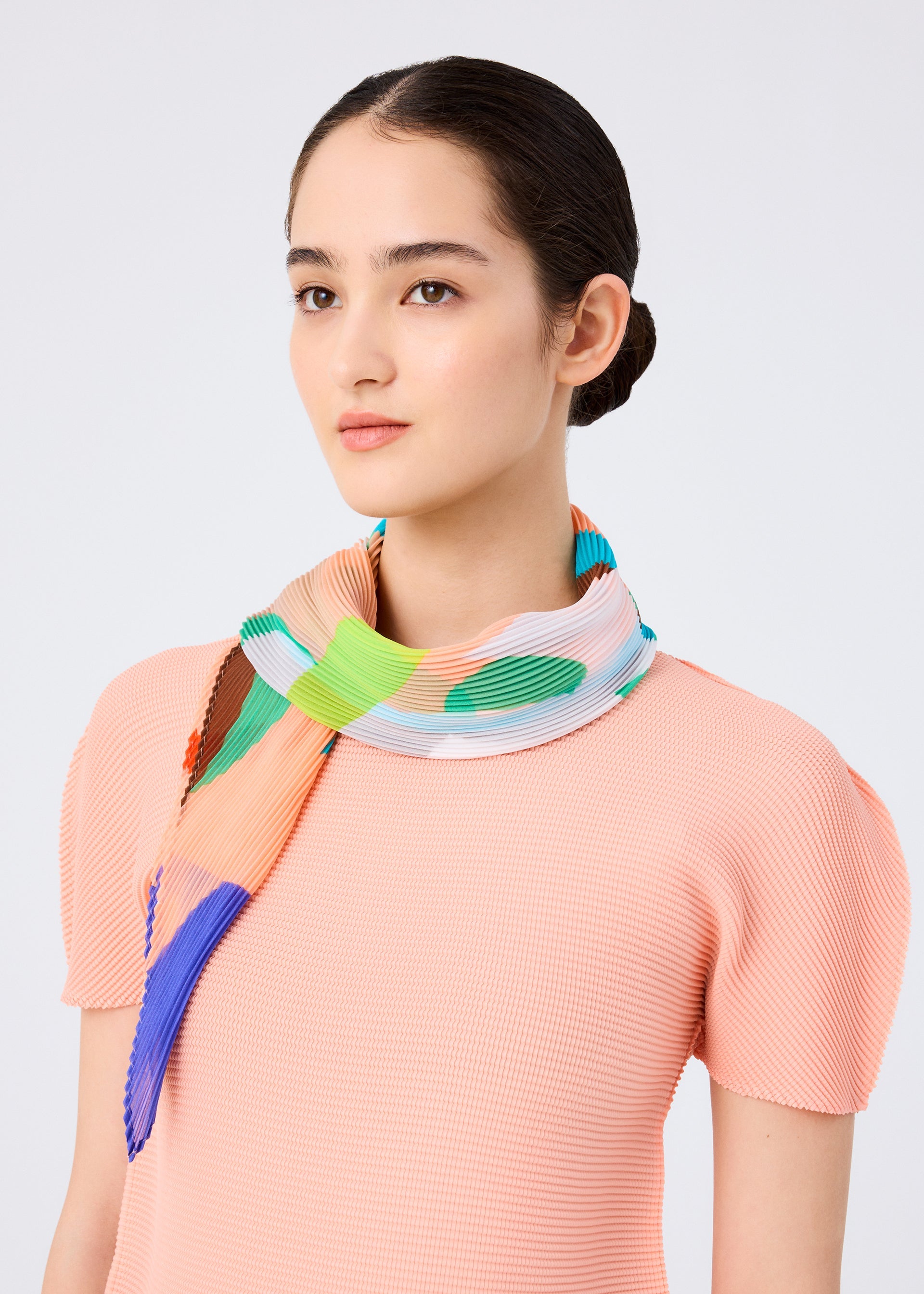 Zoomed view A woman with dark hair in a low bun wears a light pink textured top and the me ISSEY MIYAKE SURPRISE BOX PLEATS STOLE, showcasing its unique green, blue, and orange pattern against a plain light background.