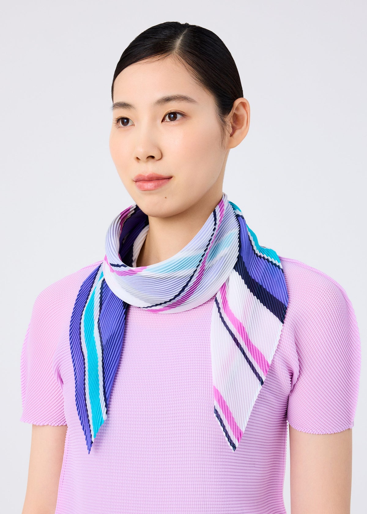 A woman with straight dark hair wears a light purple top and the SHIRT STRIPE PLEATS STOLE by me ISSEY MIYAKE—featuring blue, purple, pink, and white stripes—draped around her neck, set against a plain light background. view 5 of 5
